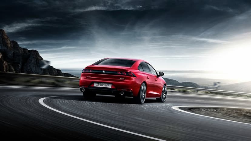 Red Peugeot 508 sedan driving through a sharp turn on a coastal mountain road under a dark cloudy sky.