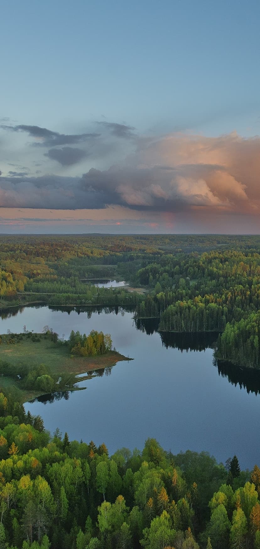Blue alpine lakes dot a vast green forest landscape beneath soft pink and purple clouds at sunset.