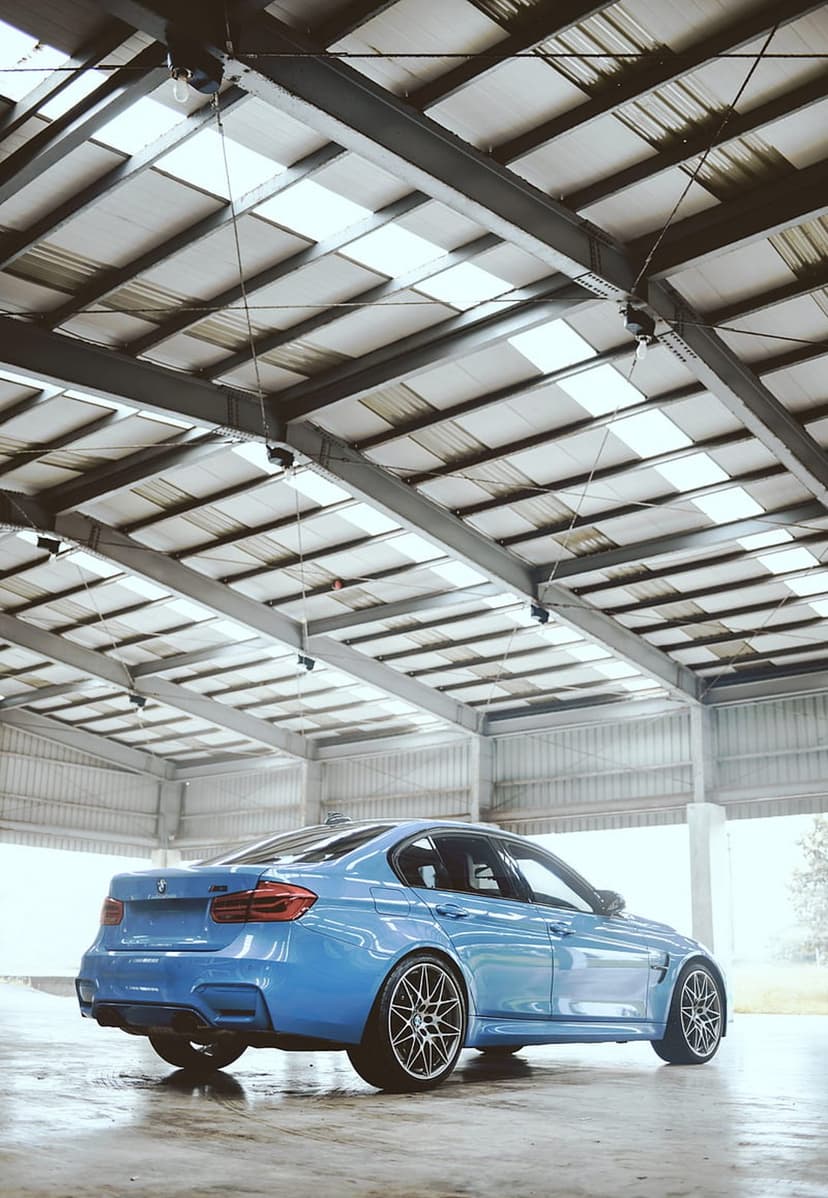 Blue BMW M3 sedan parked on a reflective wet floor inside a metal-framed industrial warehouse with bright lights.
