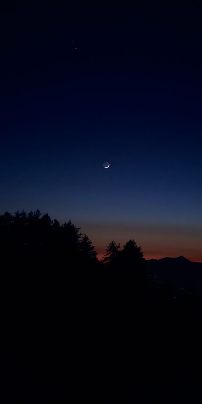 Silver crescent moon and one star above dark forest silhouettes and mountains during a blue and orange twilight.