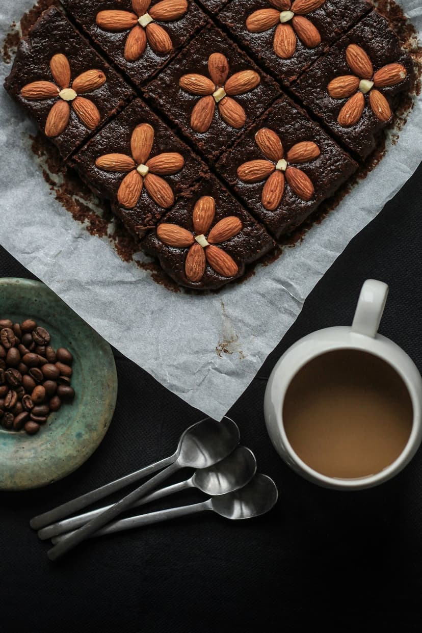 Chocolate brownies topped with almond petals sit beside a steaming coffee mug and roasted beans on parchment.
