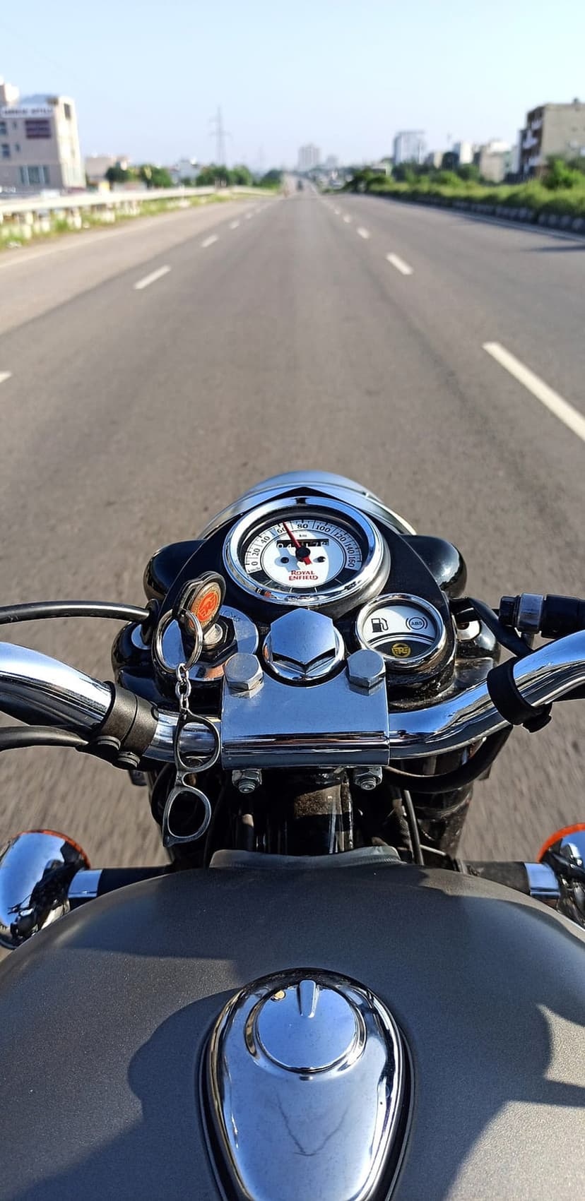 Chrome motorcycle handlebars and analog speedometer overlook a sunlit asphalt highway under a clear blue sky.