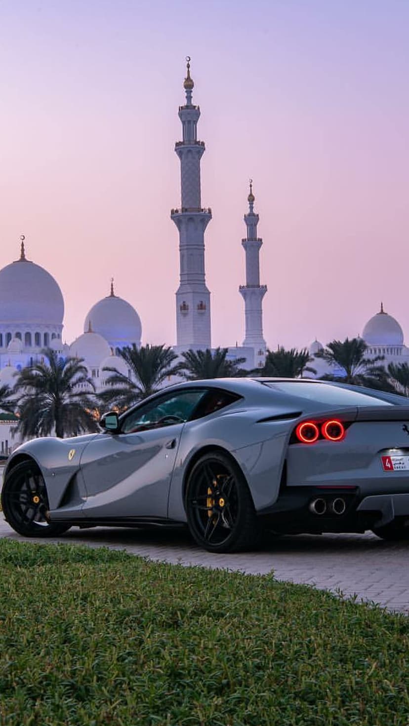 Gray Ferrari sports car parked at the Sheikh Zayed Grand Mosque with white domes and minarets under a twilight sky.