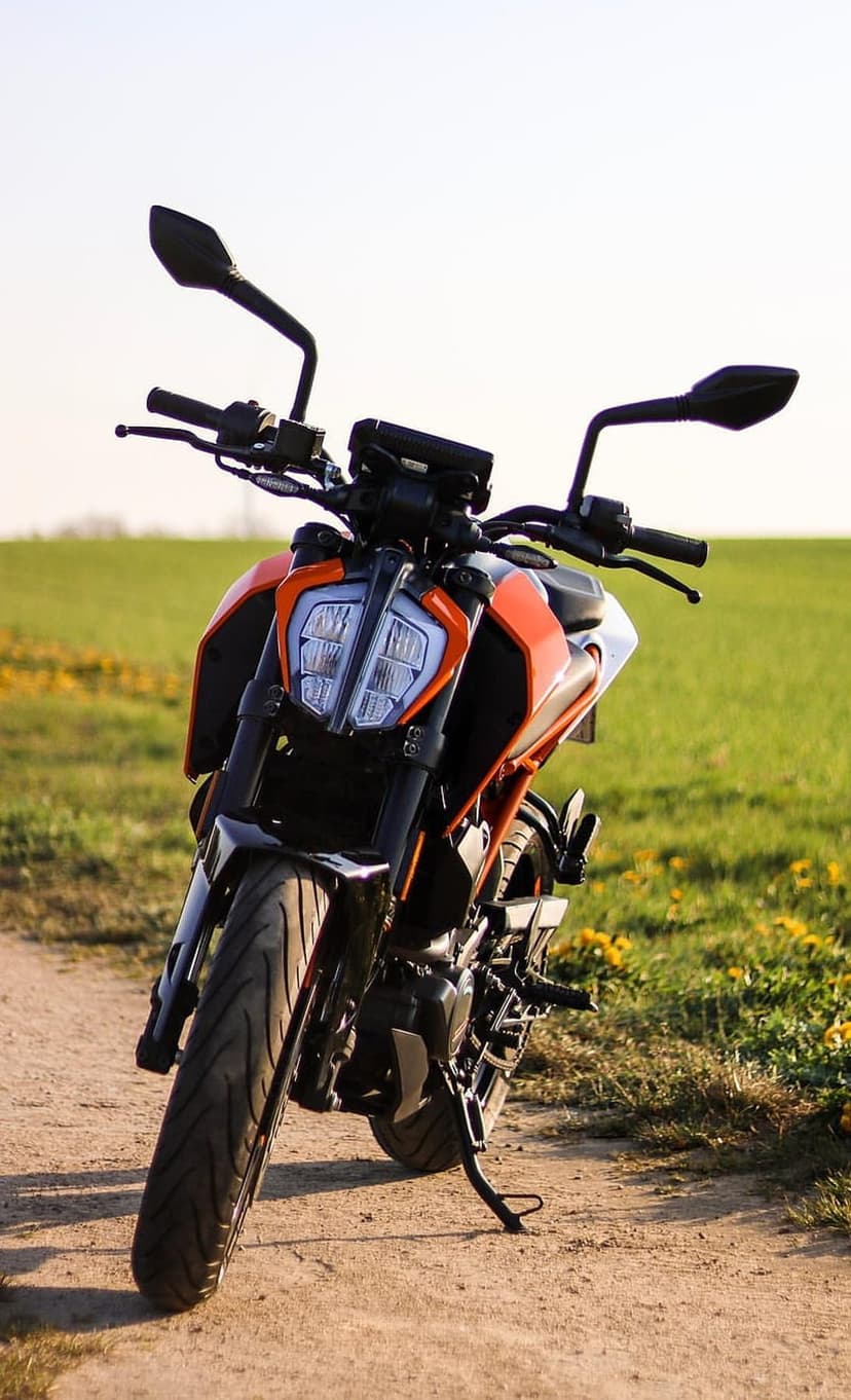 Orange KTM motorcycle with sharp fairings parked on a dirt trail near a field of green grass and yellow flowers.