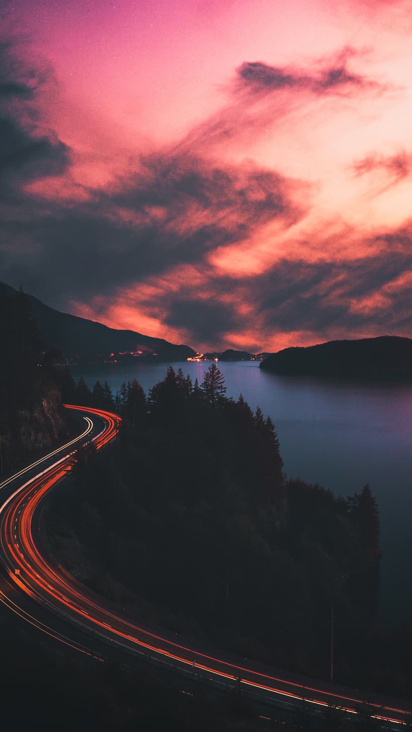 Red and white vehicle light trails streak along a winding forest road next to a calm lake under a twilight sky.