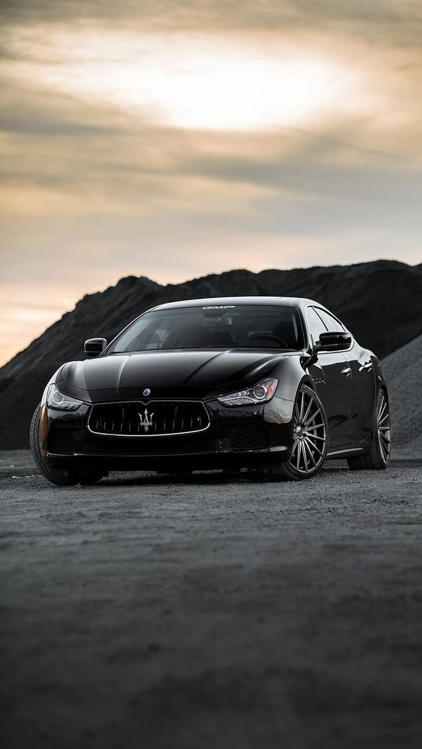 Black Maserati Ghibli sedan parked on a gravel road with mountains under a moody sky in a low-angle shot.