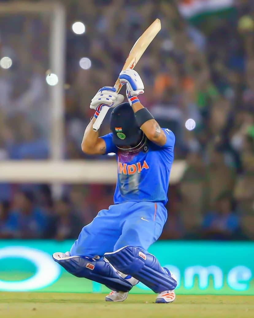 Virat Kohli swings a wooden cricket bat while wearing a blue Indian jersey against a blurred stadium background.
