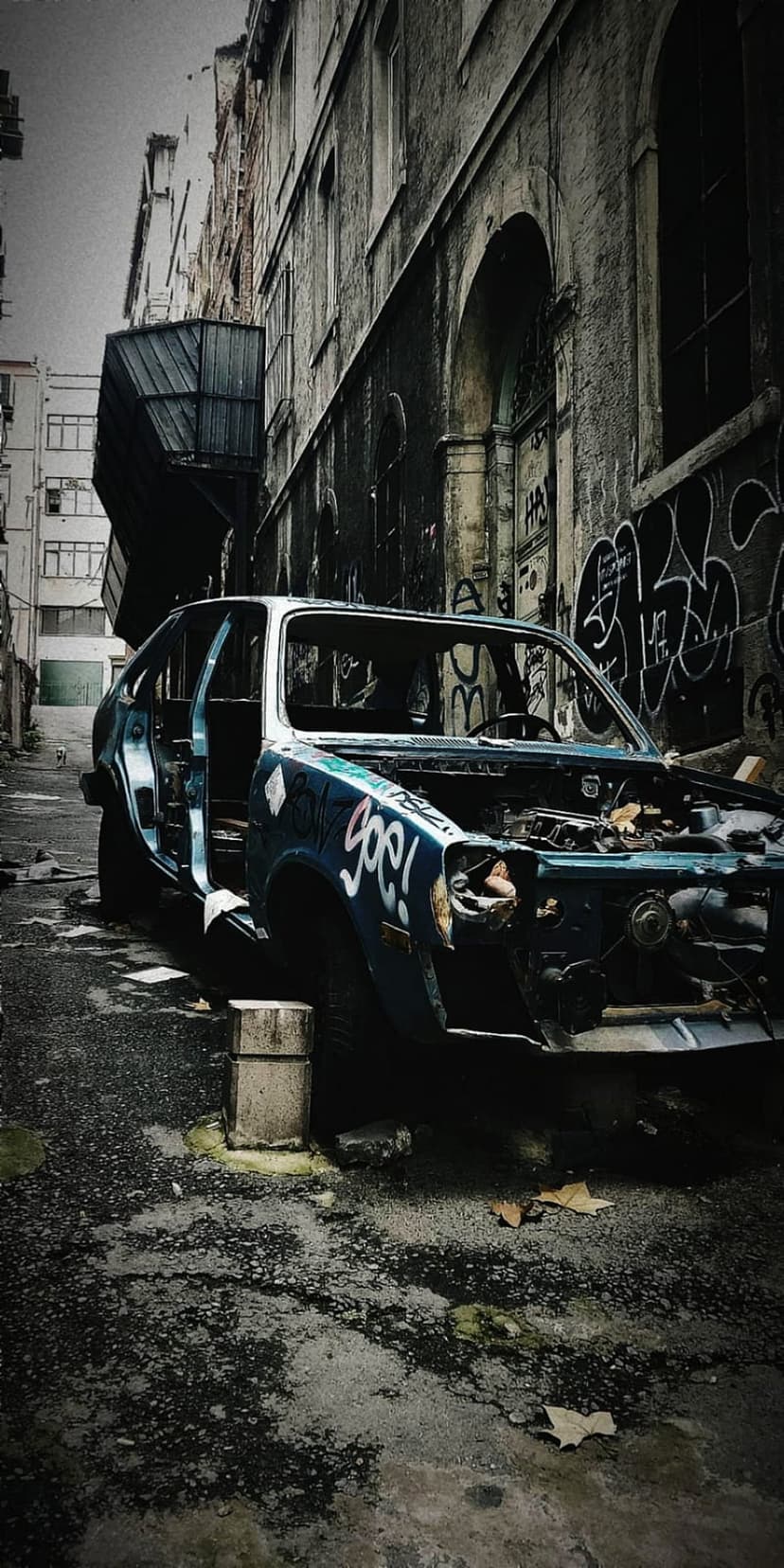 Stripped car frame in a narrow alley with colorful graffiti on brick walls and wet pavement reflections.
