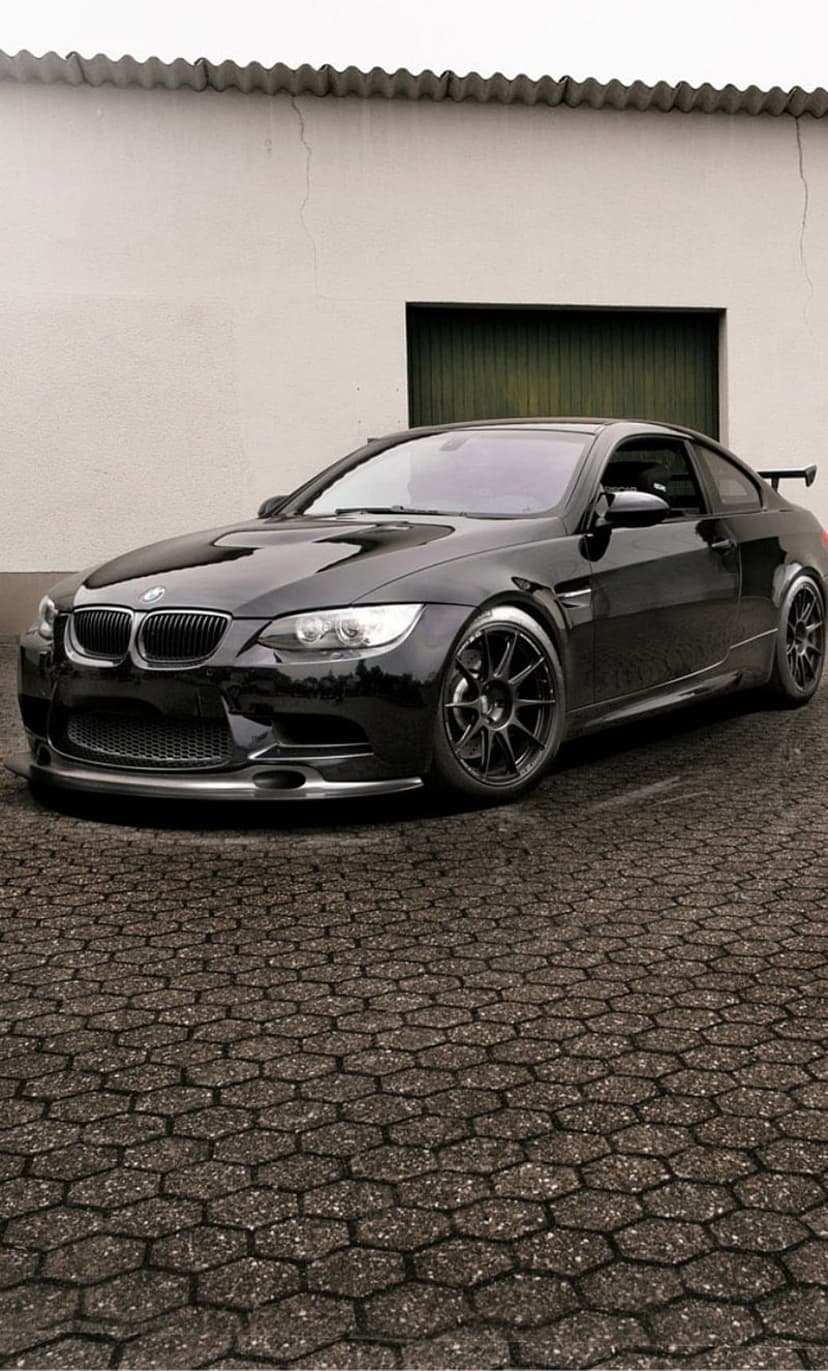 Black BMW M3 coupe with a rear spoiler and lowered stance on a cobblestone driveway near an industrial building.