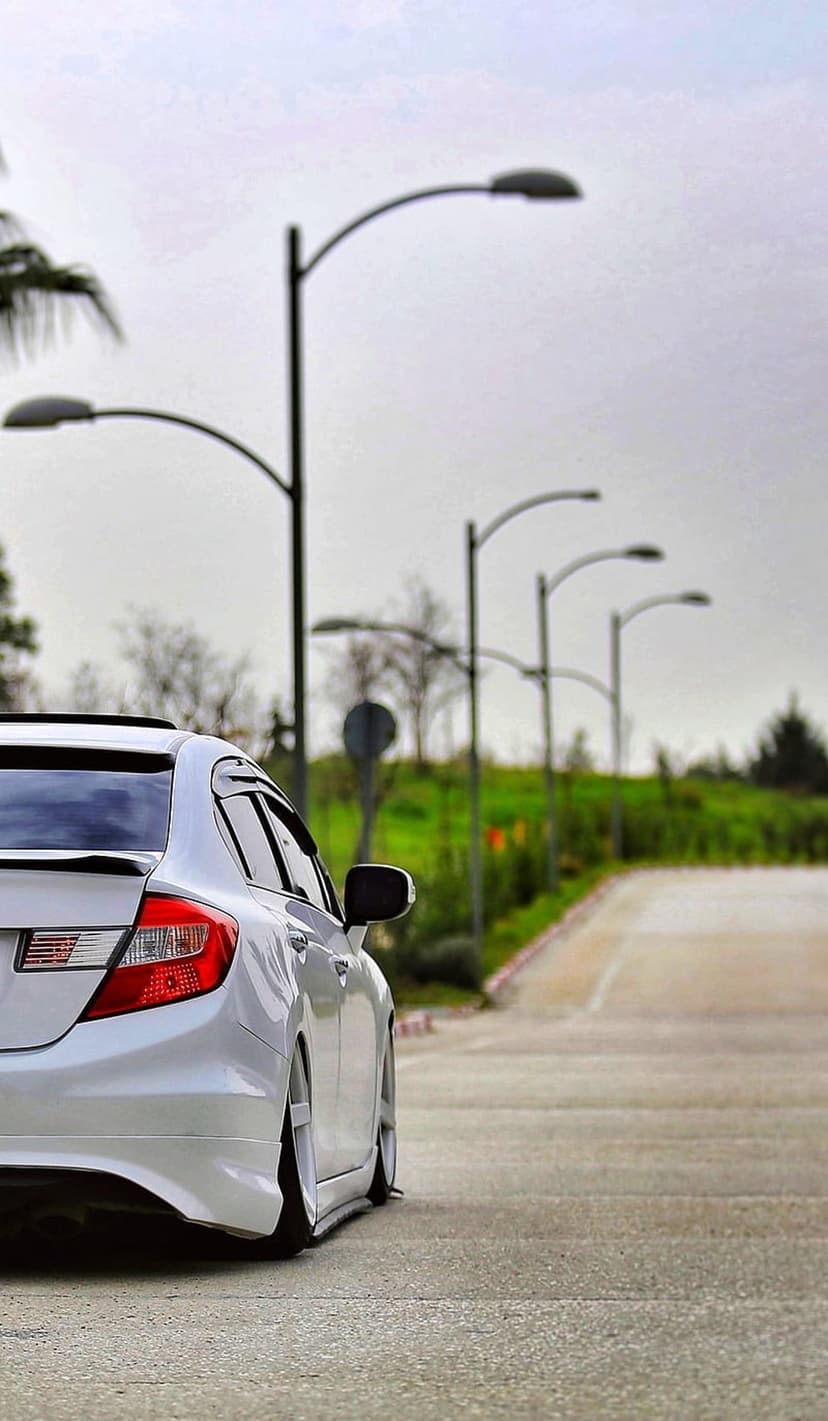 White sedan with a lowered suspension parked on a paved road beside tall streetlights and lush green trees.