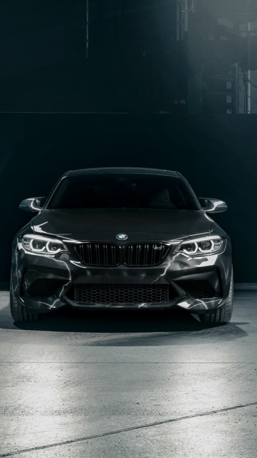 Front view of a dark grey BMW M2 with glowing white LED headlights in a dimly lit studio setting.