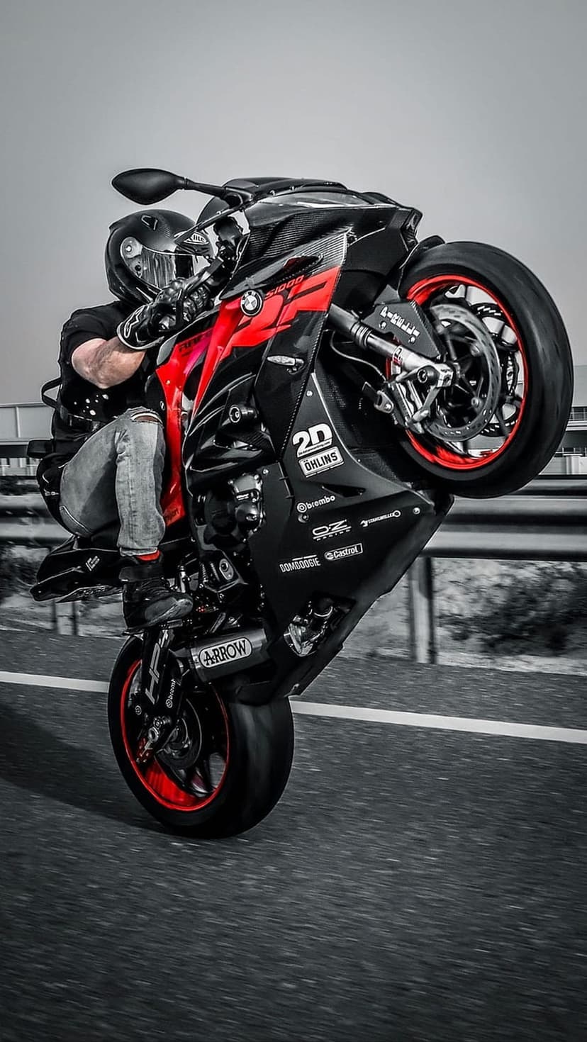 Motorcyclist in racing gear performs a high wheelie on a red and black sportbike across a gray asphalt road surface.