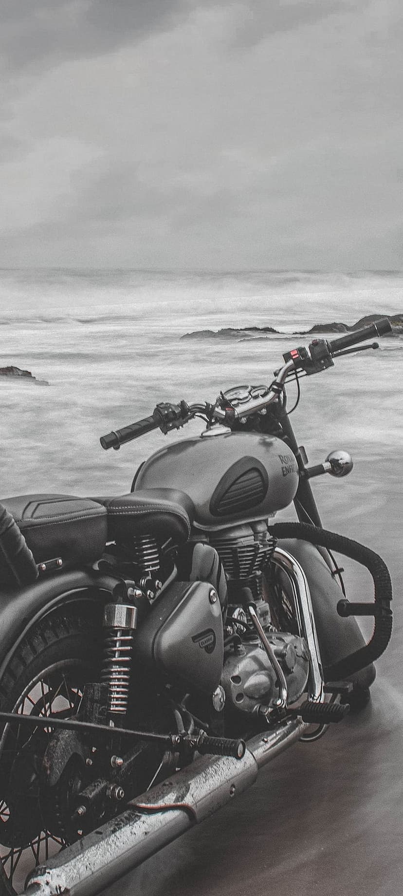 Vintage Royal Enfield motorcycle parked on wet sand with misty ocean waves crashing in a monochrome composition.