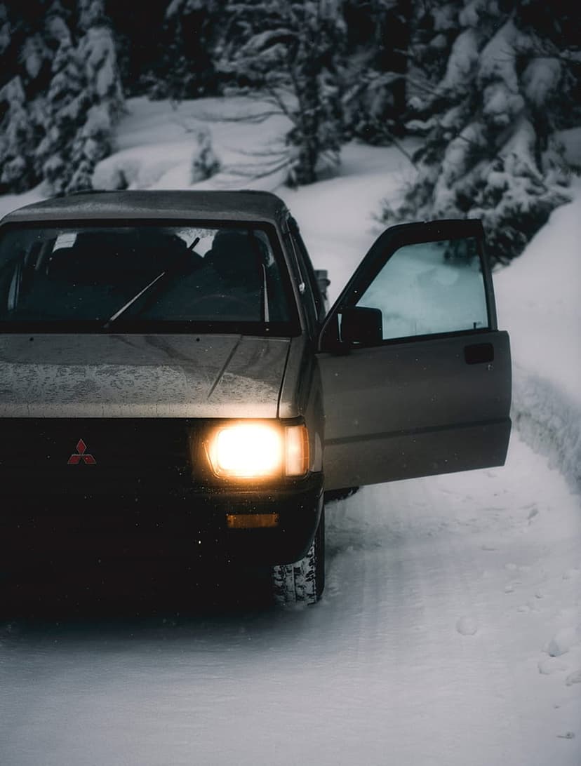 Mitsubishi pickup truck with glowing headlights parked on a snowy forest trail with an open driver side door.