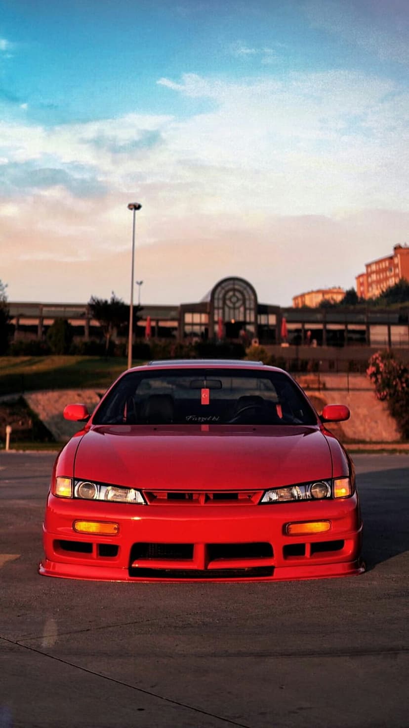 Red Nissan Silvia S14 with sharp headlights parked on asphalt against a soft twilight sky and urban architecture.