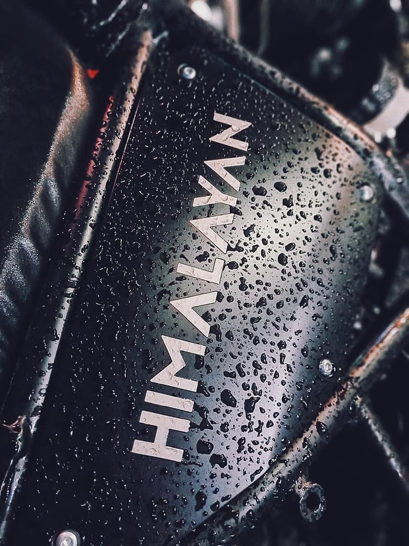 Himalayan motorcycle logo on a dark textured surface covered in clear water droplets after a rain storm.