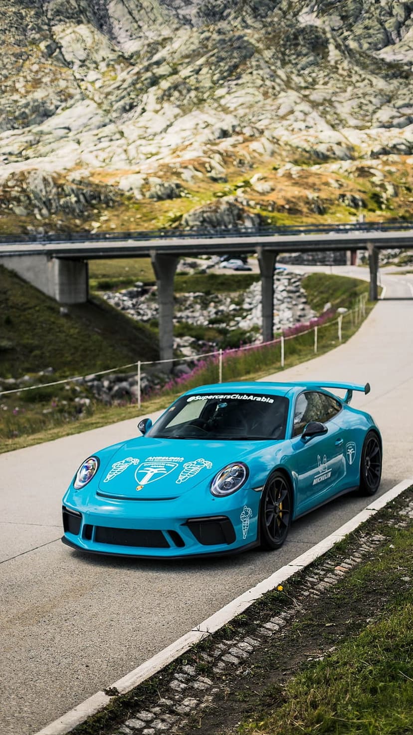 Blue Porsche GT3 with white racing decals drives across a mountain bridge surrounded by rocky cliffs.