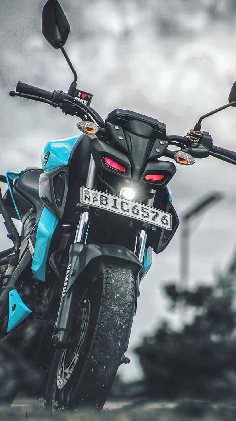 Yamaha MT-15 motorcycle with cyan bodywork and glowing red LED headlights parked against a blurred forest backdrop.