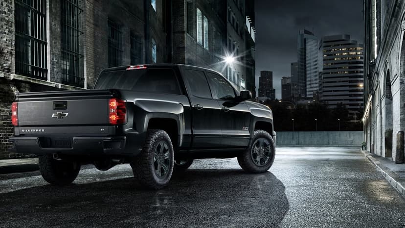 Rear view of a black Chevrolet Silverado truck parked in a dark, wet urban alley with city skyline at night.
