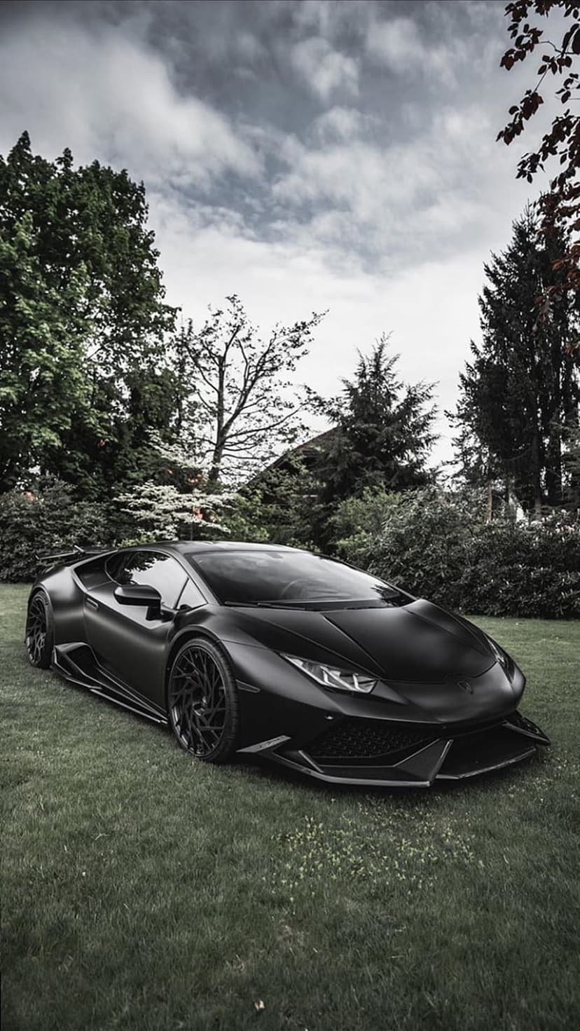 Matte black Lamborghini Huracan parked on a bright green lawn with trees and a cloudy sky in the background.