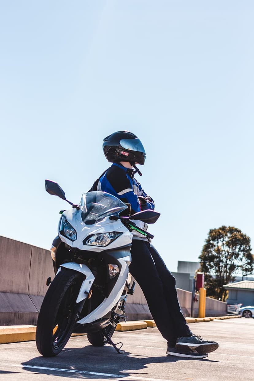 Motorcyclist in a black helmet and blue jacket leans on a white sportbike under a bright blue summer sky.