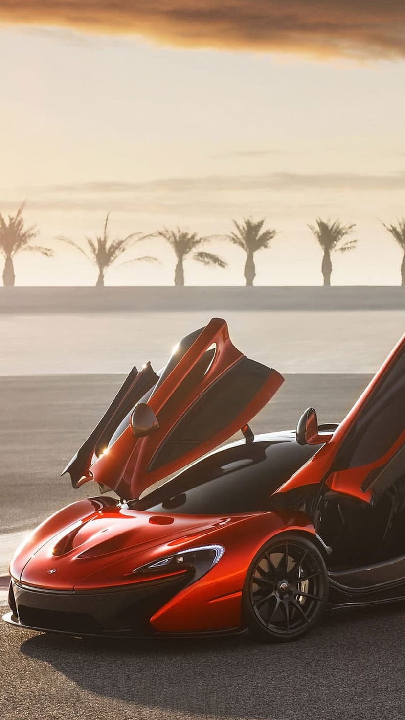 Red McLaren P1 supercar with open dihedral doors parked before silhouetted palm trees during a golden sunset.