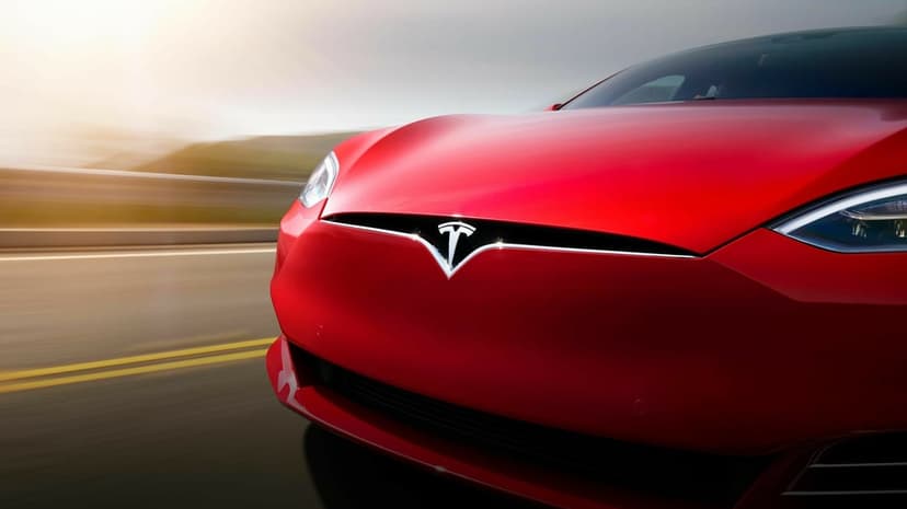 Close-up of a red Tesla front grille and emblem with motion blur on a highway under warm sunlight.