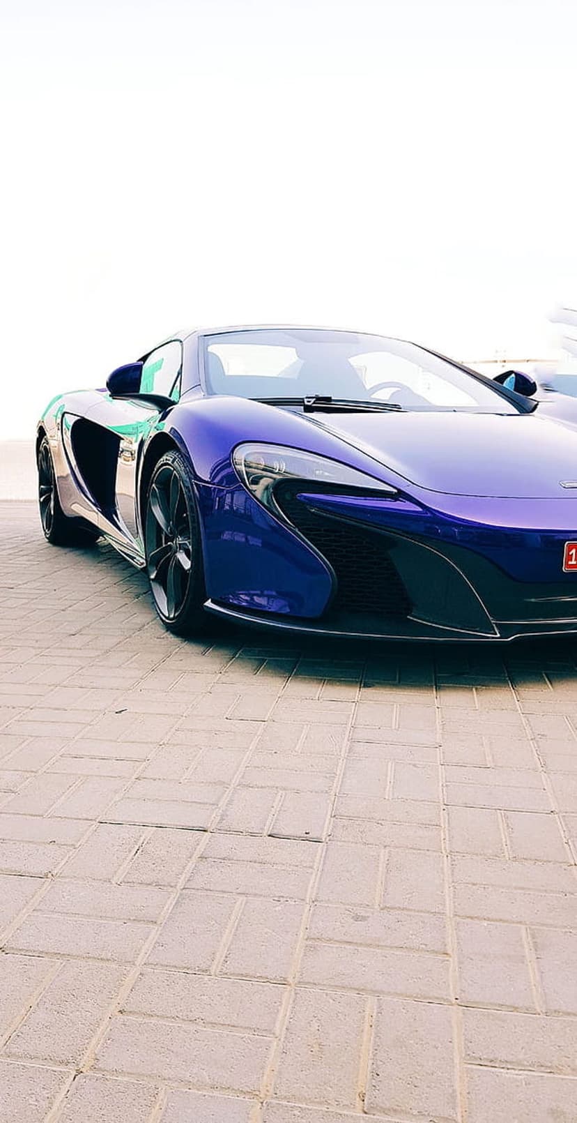 Purple McLaren sports car parked on light-colored brick pavement against a bright white minimalist background