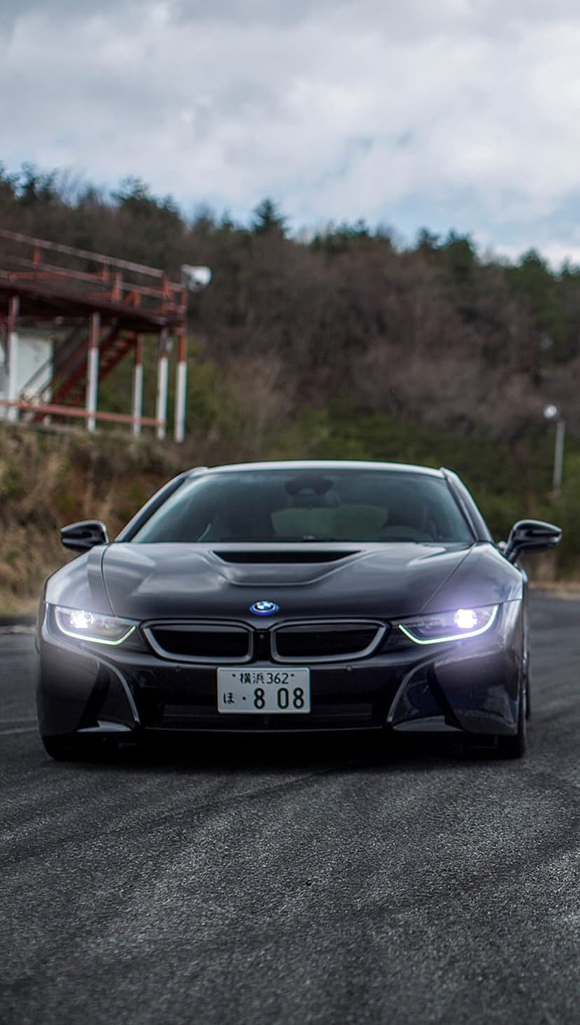 Dark BMW i8 sports car driving head-on through a curve with glowing headlights under a gloomy overcast sky.