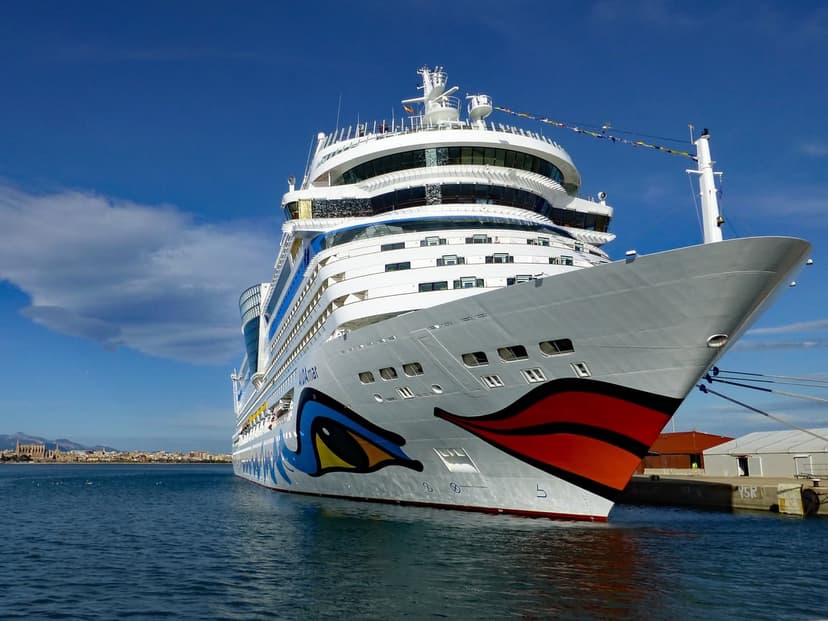 AIDAstella cruise ship with red lips livery docked at a harbor under a bright blue sky with wispy clouds.