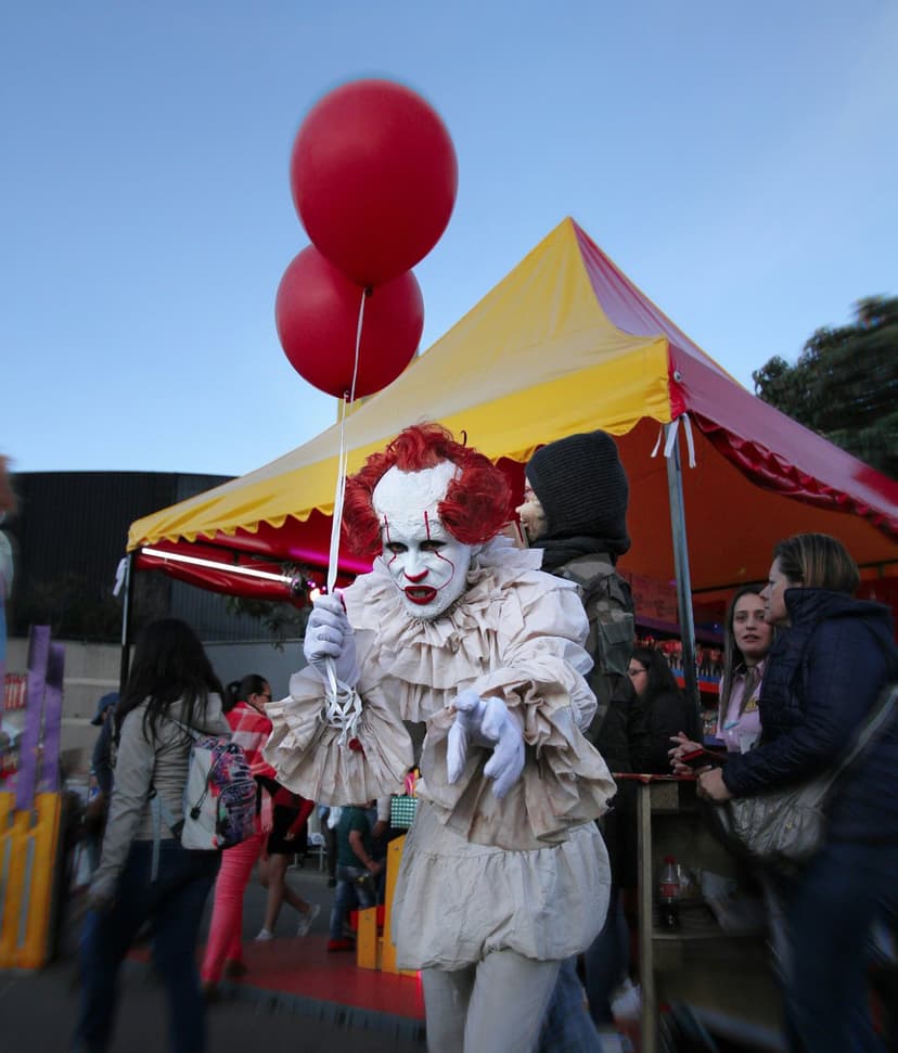 Frightening clown with white face paint and red balloons stands before a yellow carnival tent at an event.