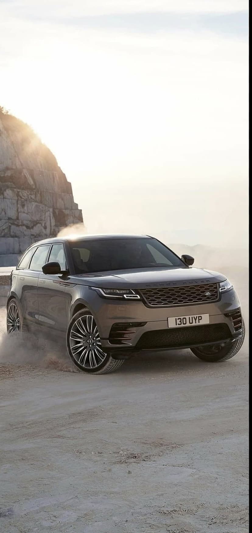 Range Rover Velar SUV driving on a dirt track, kicking up a dust cloud against a rocky cliff in hazy sunlight.