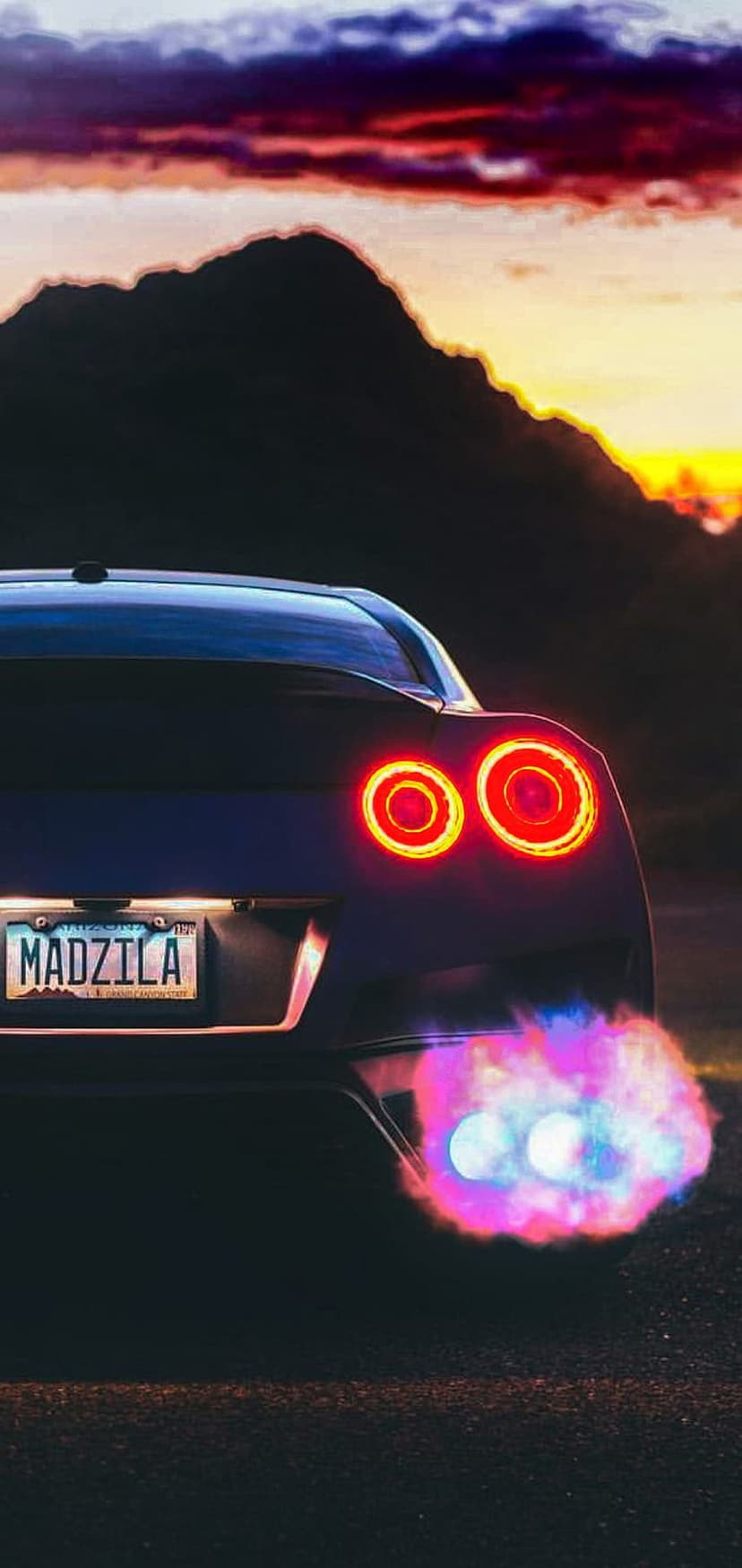 Sports car with circular taillights and colorful exhaust flames against a mountain sunset background.