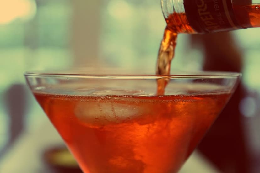 Aperol Spritz liquid pouring into a crystal martini glass filled with ice cubes against a soft golden background.
