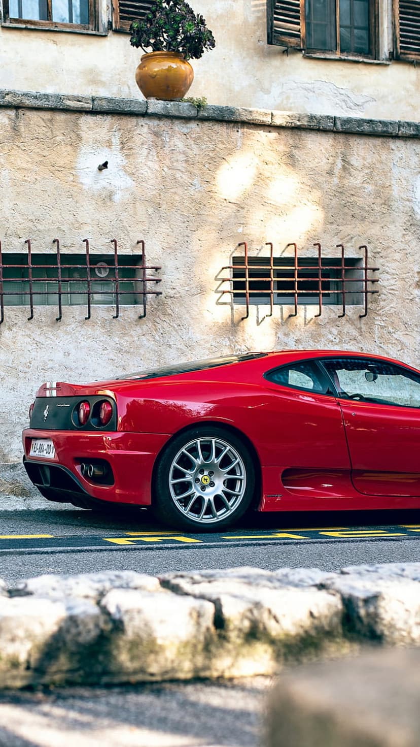 Red Ferrari sports car parked beside a weathered stone wall with window grates and a small potted plant.