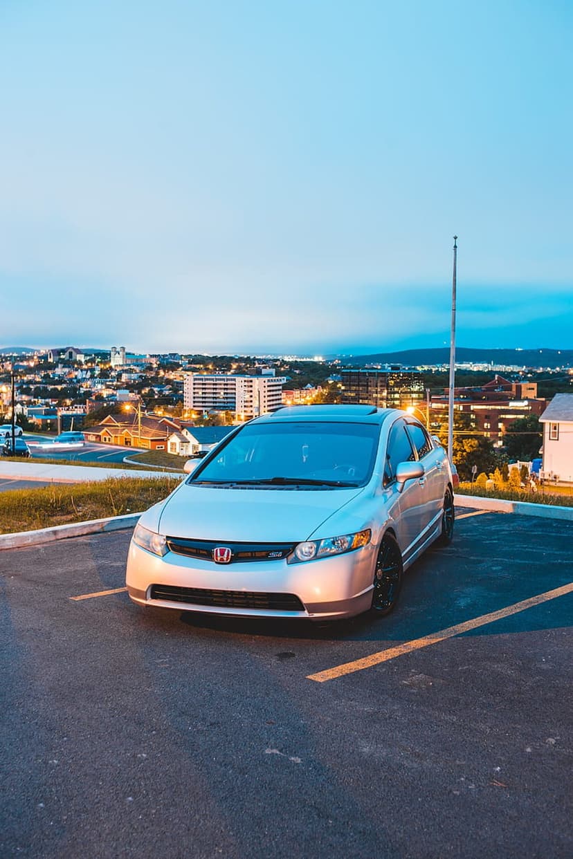 Silver Honda Civic with glowing headlights parked on a hill overlooking a twinkling city skyline at twilight.