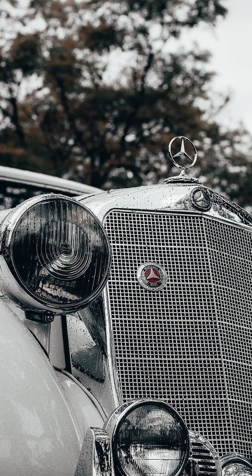Mercedes-Benz three-pointed star hood ornament and glass headlight glistening with raindrops on a chrome grille.
