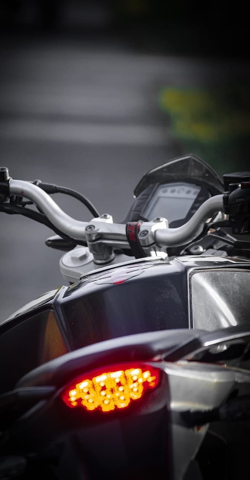 Motorcycle handlebars and a bright red LED tail light glow against a dark, blurred night street background.