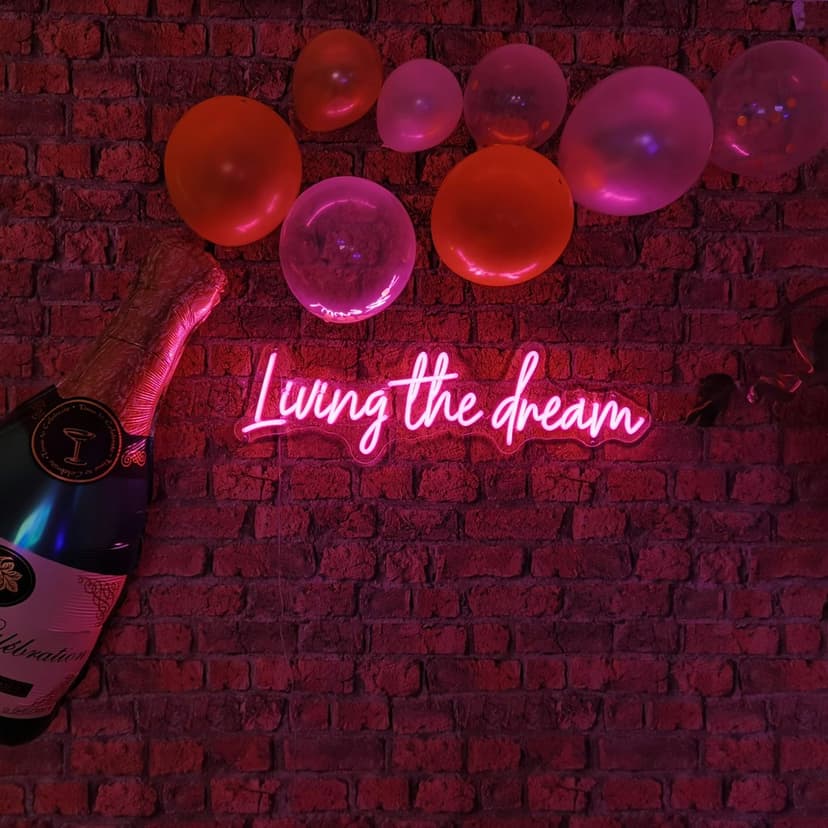 Pink neon sign reading Living the Dream glows on a brick wall next to a champagne bottle and festive balloons.