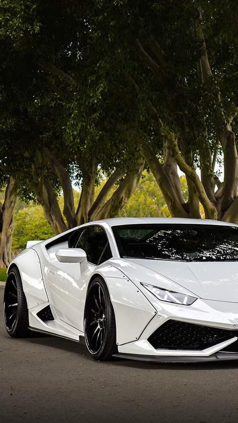 White widebody Lamborghini Huracan parked on an asphalt road beneath lush green trees with dappled sunlight.