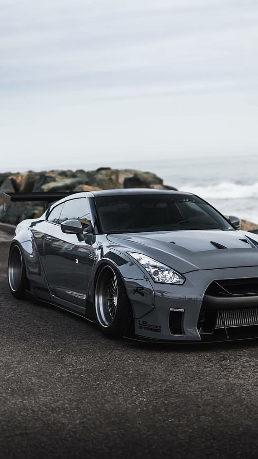 Gray Nissan GT-R with a widebody kit parked on a coastal road against a cloudy sky and rocky ocean shore.