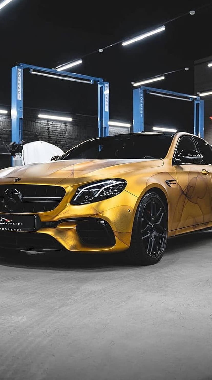 Gold Mercedes-Benz sedan with swirl patterns parked in a garage with blue lifts and bright overhead lights.