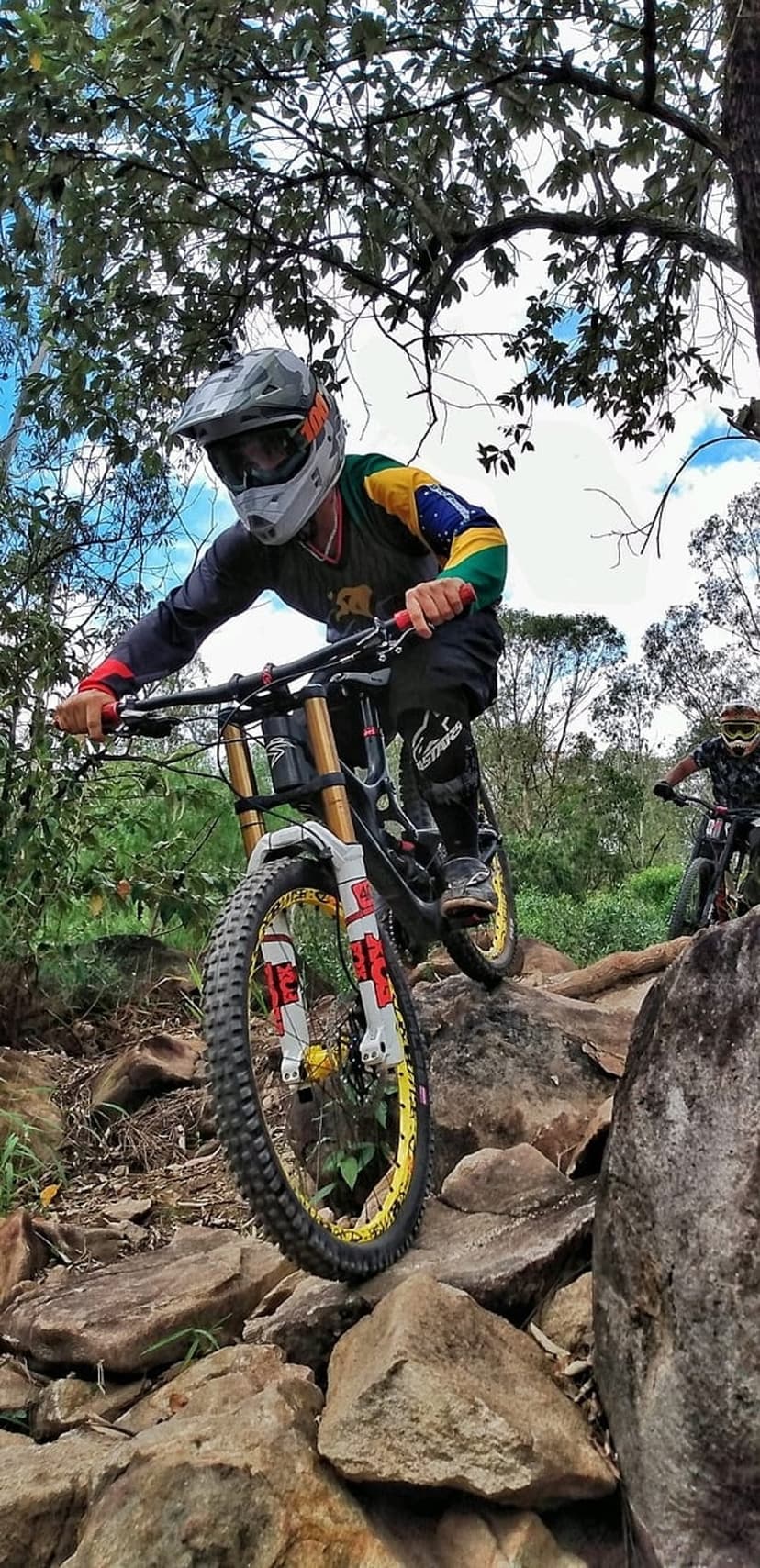 Mountain biker in a Brazilian jersey navigates a rocky downhill trail amidst lush green forest scenery.