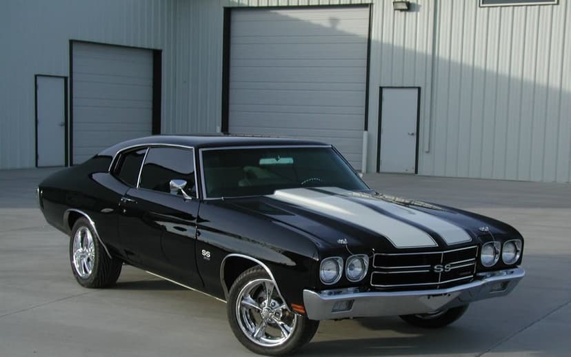 Black 1970 Chevrolet Chevelle SS with white racing stripes parked in front of a white metal industrial building.