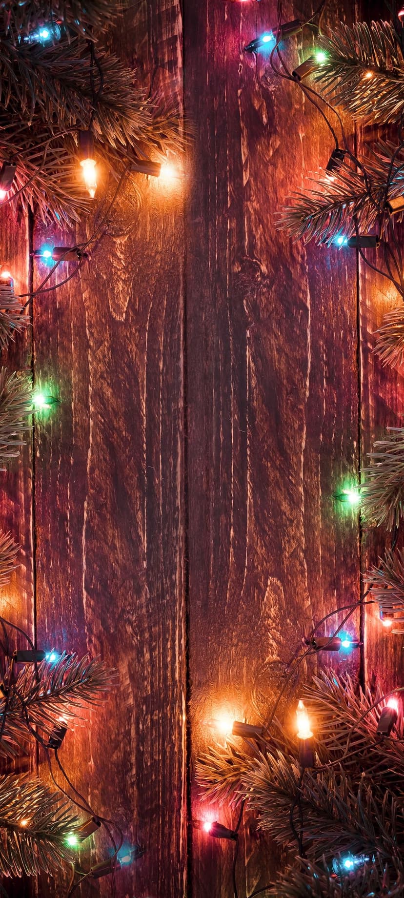 Multi-colored Christmas lights and pine needles rest on a dark, rustic wooden surface with warm glowing bokeh.