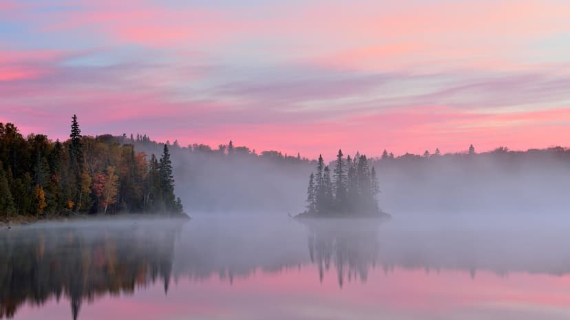 Misty lake with small tree-covered islands under a soft pink sunrise sky reflecting on the still water surface.