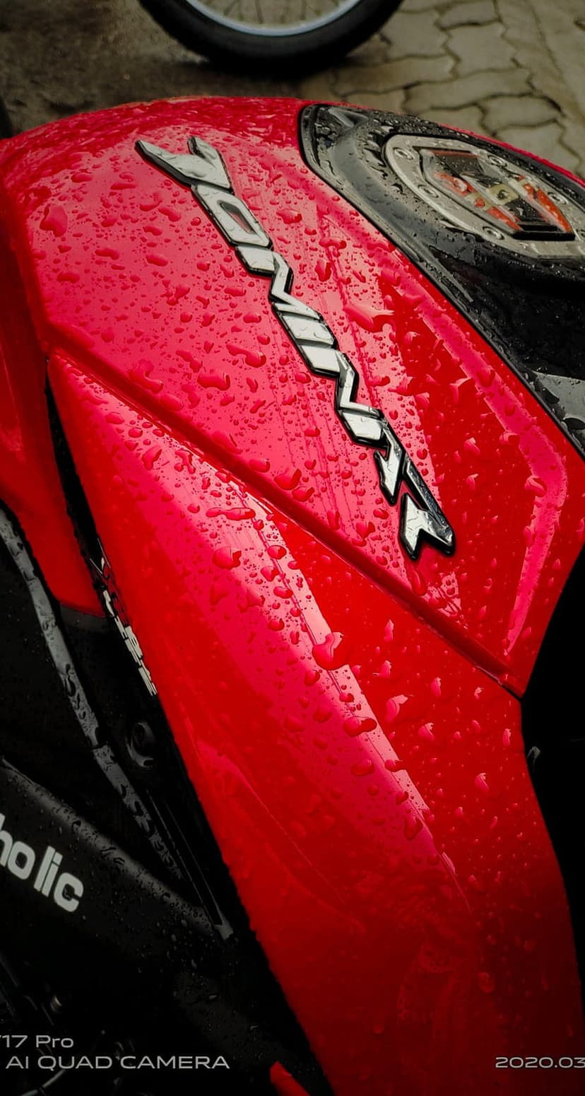 Red motorcycle fuel tank covered in water droplets with a silver chrome emblem reflecting its surroundings.