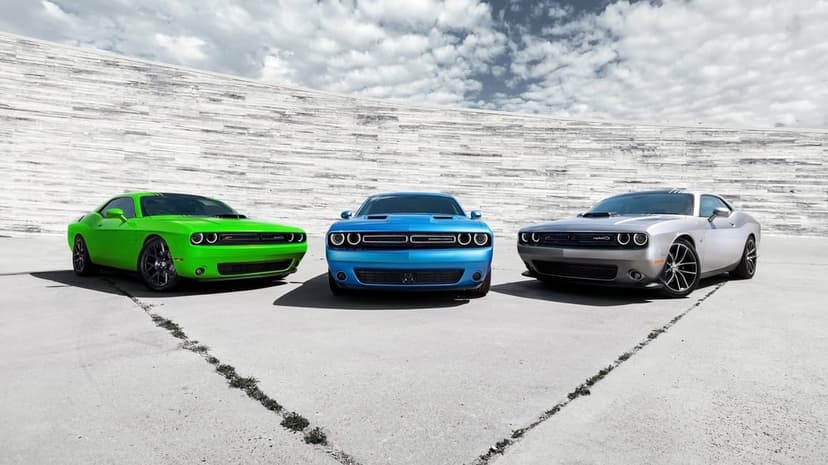 Three Dodge Challenger muscle cars in green, blue, and silver parked on concrete against a grey stone wall.