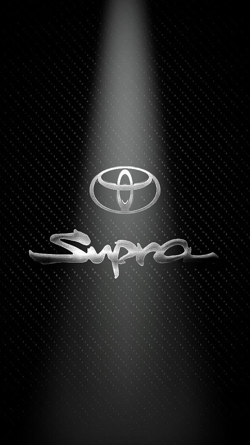 Toyota Supra chrome emblem and lettering illuminated by a spotlight on a dark textured carbon fiber background.