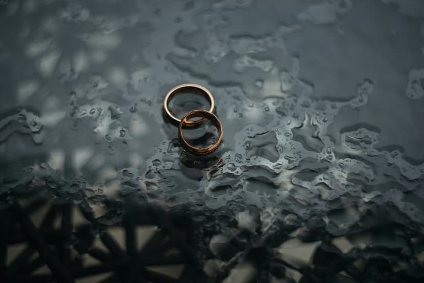Two gold wedding rings lie on a dark wet glass surface covered in small translucent water droplets and reflections.